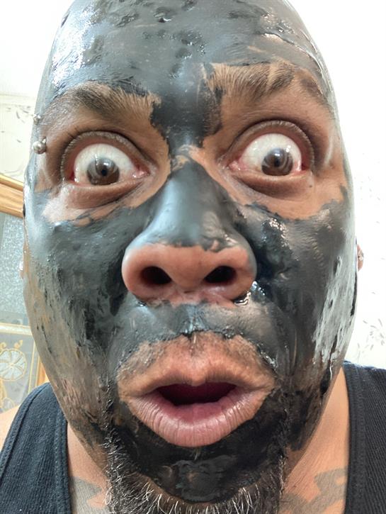 Person shows a shocked face while applying a black facial mask in a bright indoor setting.