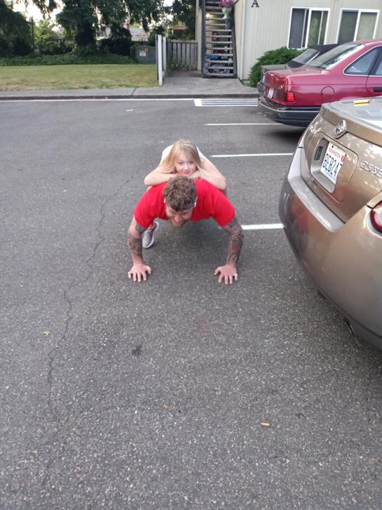 A child is playfully riding on a father's back while he performs push-ups in a parking lot.