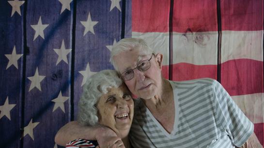 Joyful elderly couple embraces, smiling broadly in front of an American flag, showcasing their bond.
