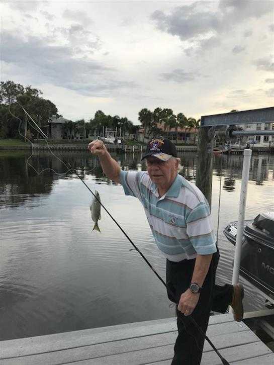 An elderly man stands on a dock, excitedly holding up a fish he just caught from the river.