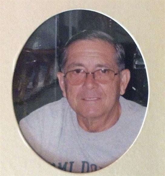An elderly man with glasses smiles gently while seated indoors, exuding warmth and kindness.
