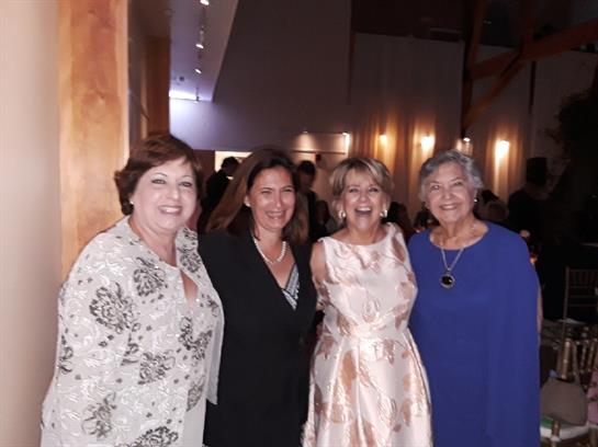 Four women smile and pose together at a lively indoor event in the evening, showcasing their attire.