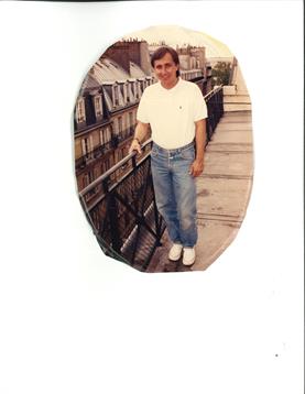 A person relaxes on a balcony in jeans and a T-shirt, enjoying a sunny city view.