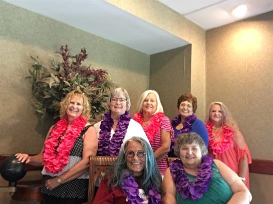 Eight women in leis joyfully celebrate a special occasion indoors with floral decor.