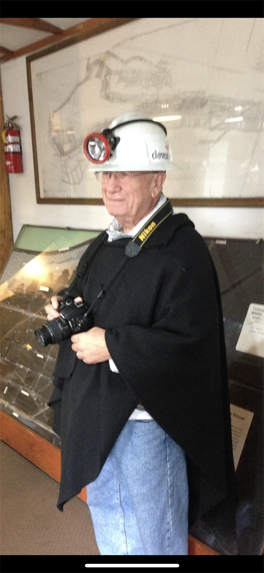 A man in a mining helmet and poncho stands proudly while holding a camera inside a mining exhibit.