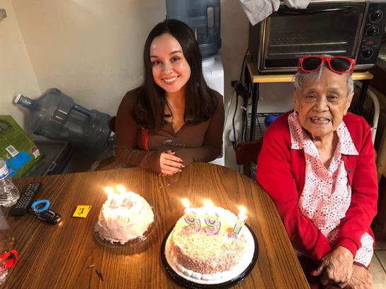 Two women smile at a table with birthday cakes, candles lit, in a cozy indoor setting.