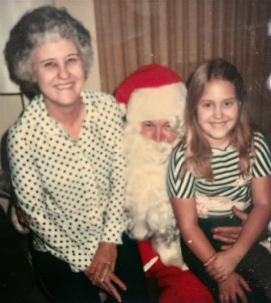 A young girl and her grandmother happily pose with Santa Claus in a festive indoor setting.