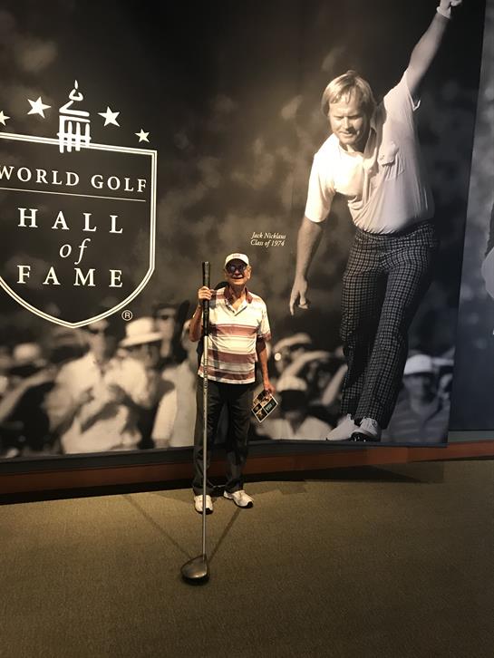 A person proudly stands next to a large golf display at the Hall of Fame, celebrating legends.