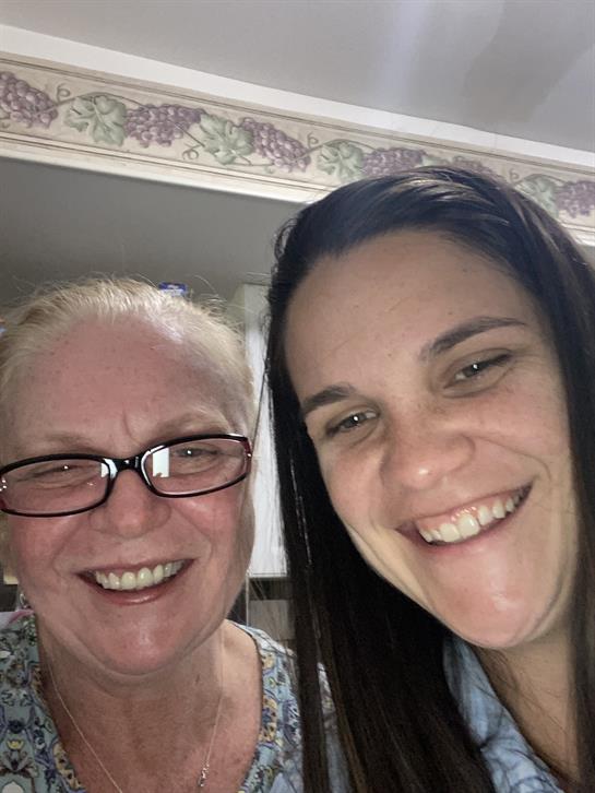 Two women pose closely, smiling widely, enjoying each other's company in a warm, home environment.