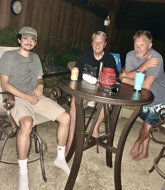 Three friends sit around a table in a backyard, sharing drinks and enjoying a casual evening.