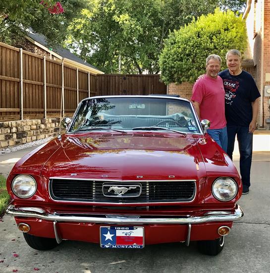 Two men pose beside a vintage red Mustang parked in a quiet neighborhood during daylight.