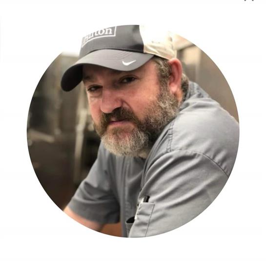 A man with a beard wearing a cap appears deep in thought while working in a workshop.