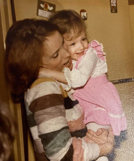 A joyful mother holds her smiling daughter close in a warm indoor setting, surrounded by love.