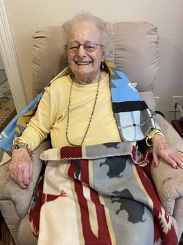 An elderly woman smiles happily, wrapped in colorful blankets as she relaxes in a chair.