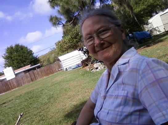 An elderly woman smiles in a garden, sitting comfortably on a chair under the sun.