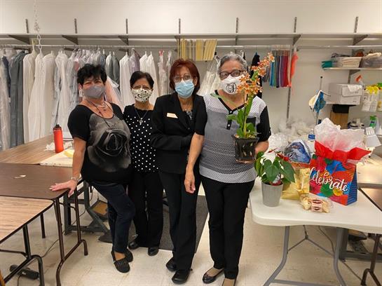 Four women wearing masks gather around a table with a potted plant in a work environment.
