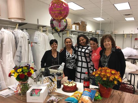 A group of five friends gathers in a bright kitchen, celebrating with a cake, snacks, and balloons.