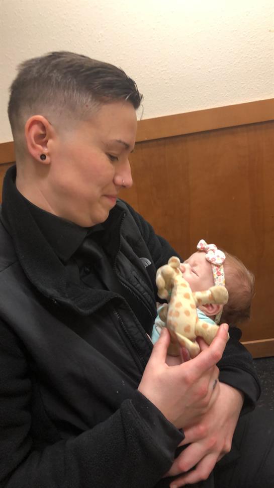 A caregiver holds a baby, sharing smiles and playing with a soft toy in a cozy indoor space.