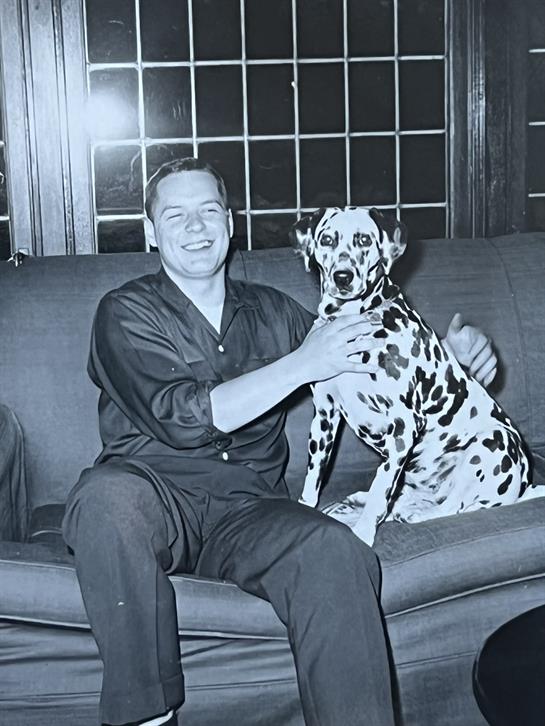 A man casually dressed interacts with a Dalmatian on a couch during a cozy evening indoors.