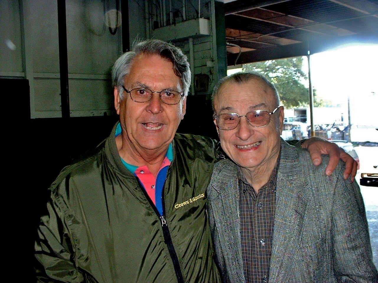 Two men stand next to each other with smiles on their faces while talking in a building.