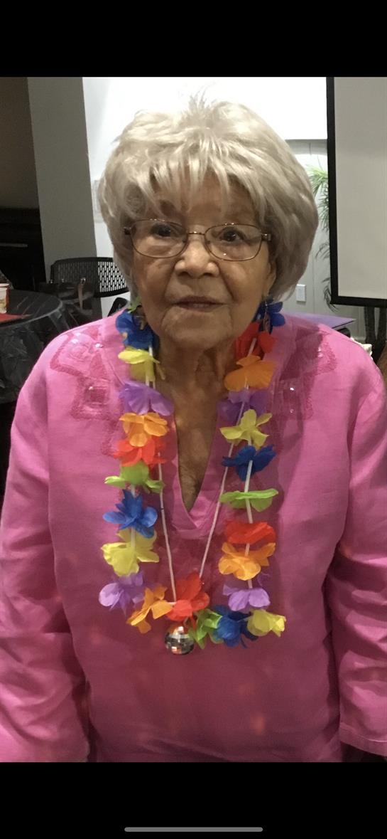 Elderly woman looks happy while wearing a colorful lei and enjoying a festive celebration indoors.