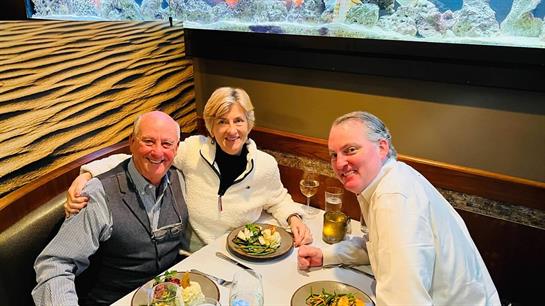 Three diners smile and share a meal at a well-decorated table near a large aquarium.