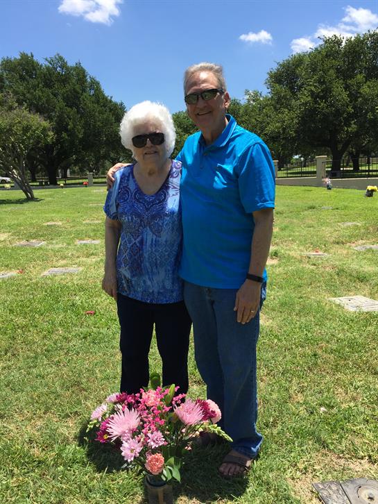Two family members gather in a cemetery, standing beside a flower arrangement.