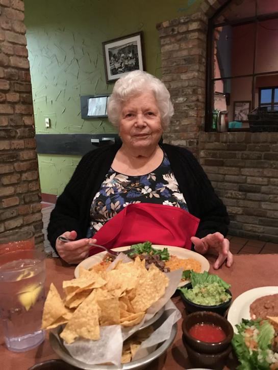 Elderly woman sits at a table with a plate of nachos and guacamole, smiling contentedly.