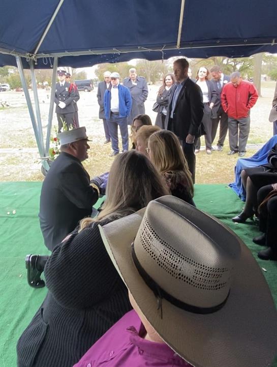 A group gathers under a tent for a ceremony, with some sitting and others standing in attendance.