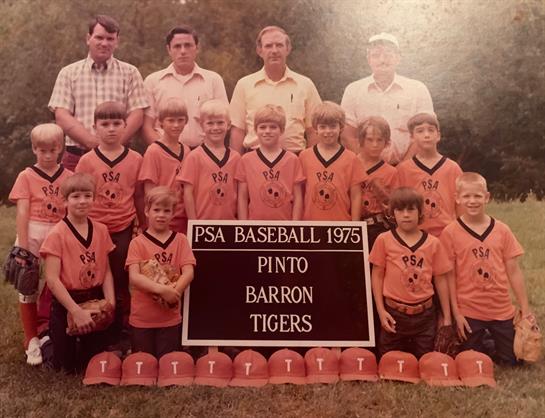 A youth baseball team in orange jerseys stands proudly with their coaches at a local field.