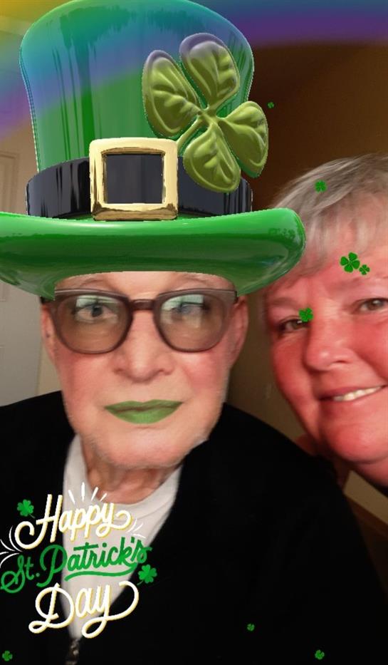 Two seniors take a joyful selfie wearing St. Patrick's Day hats and themed decorations.