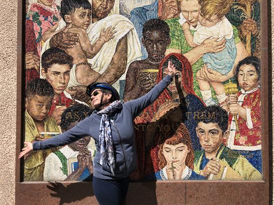 A person poses joyfully next to a vibrant mural depicting various cultural faces and scenes.