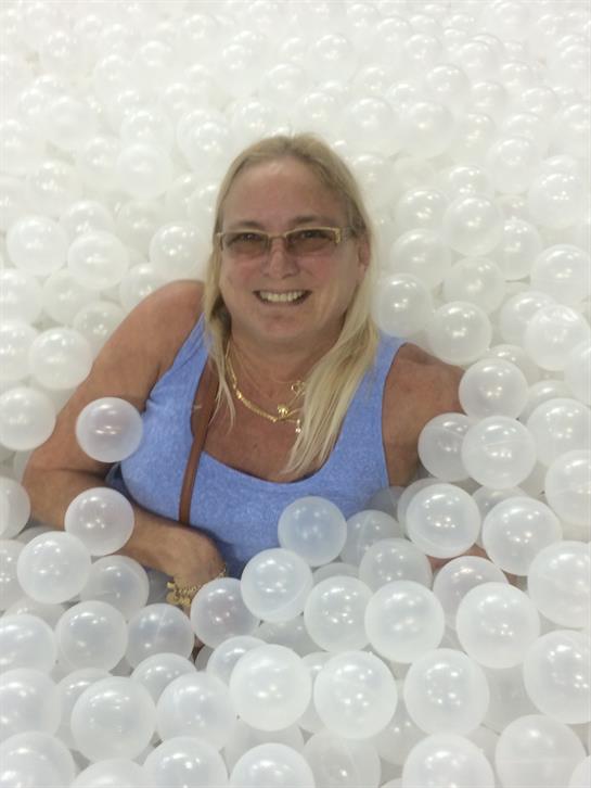 A woman with blonde hair smiles joyfully while lounging in a ball pit filled with clear balls.