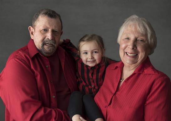 Grandparents smile proudly while holding their granddaughter in a warm indoor environment.