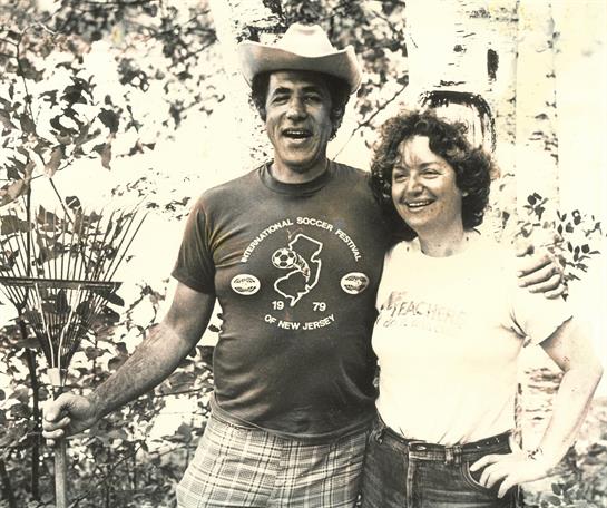 A cheerful couple poses together in a lush garden, both smiling and enjoying the sunny weather.