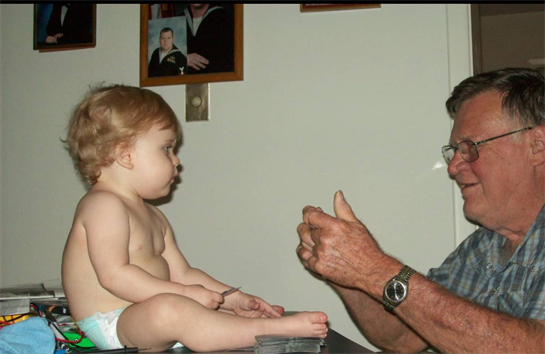 An elderly man engages with a toddler, who sits on a table, showcasing a warm family interaction.