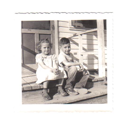 Two children relax on the porch steps of a rustic home under warm sunlight, smiling.