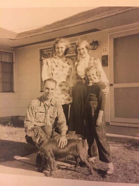 Three people pose happily outside their home with a dog on a sunny afternoon.