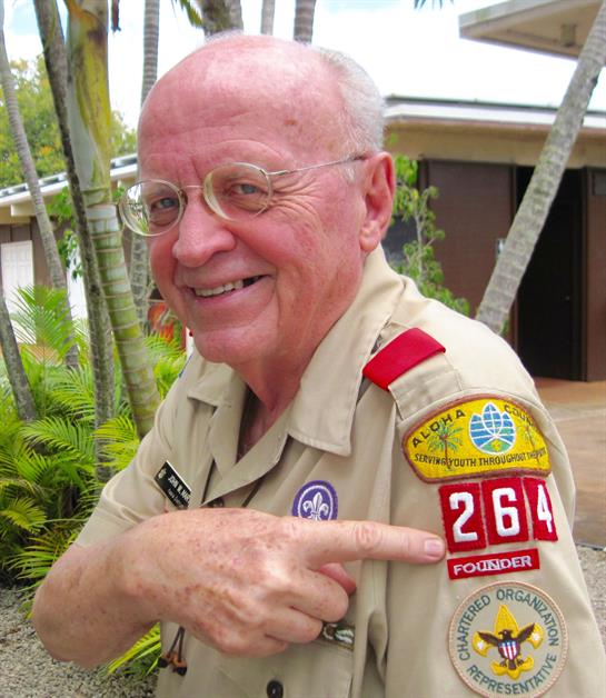 Elderly man proudly points to his scout uniform patches in a vibrant garden during daytime.