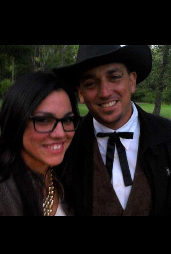 Two individuals smile while dressed elegantly, enjoying a warm day outdoors surrounded by greenery.