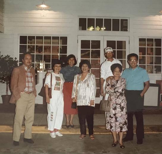 A group stands outside a restaurant during evening hours, smiling and dressed in casual attire.