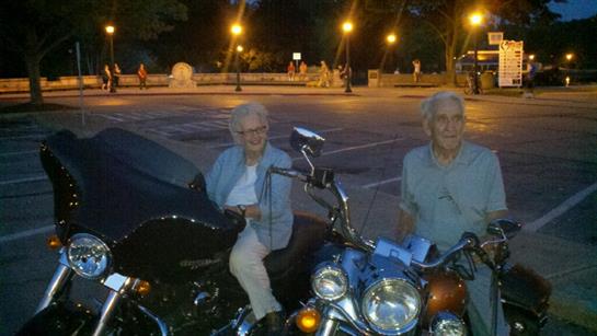 Two older individuals sit on their motorcycles, smiling in a vibrant park at night.