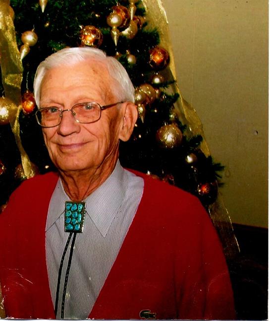 An elderly man stands smiling close to a Christmas tree adorned with ornaments and lights.