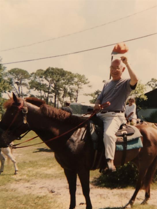 A man rides a brown horse while waving a hat at spectators at a community gathering.