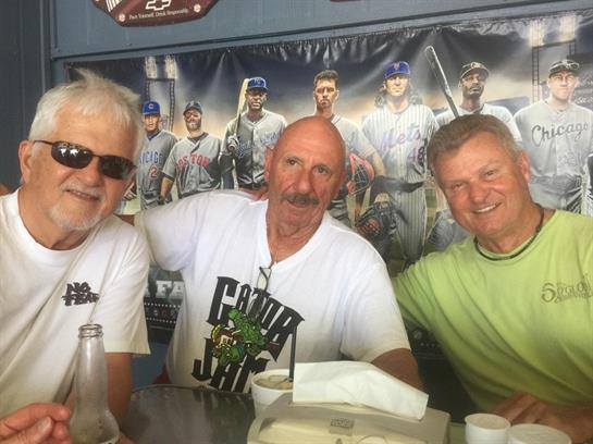 Three men smile together at a table in a sports bar, surrounded by baseball memorabilia.