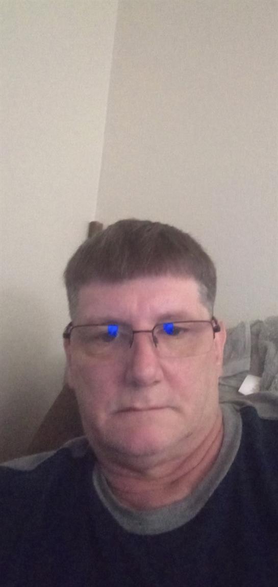 Man with glasses and short hair sits indoors, gazing at the camera with a calm expression.