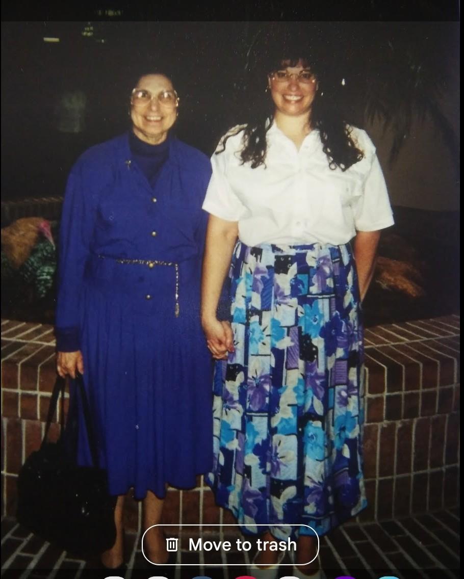 Women stand close, smiling while dressed in vibrant clothing, in a cozy indoor environment.