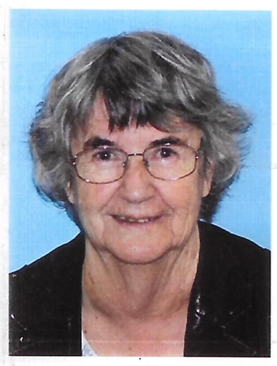 A senior woman with curly grey hair and glasses smiles gently, highlighting her warmth and kindness.