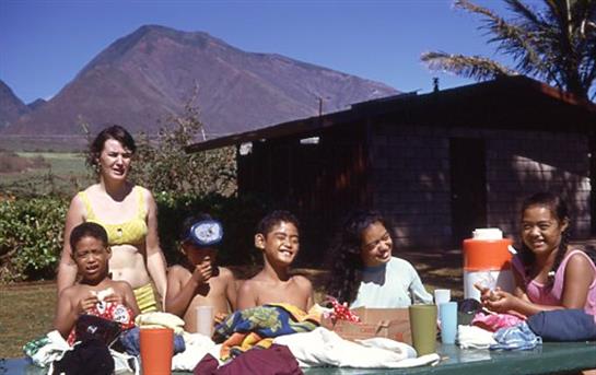 Children and adults celebrate outdoors, surrounded by mountains, dressed for a warm, sunny day.