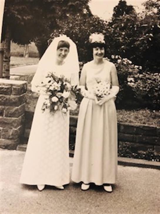 Two women in a garden: one in a wedding gown and the other in a long dress, both with bouquets.